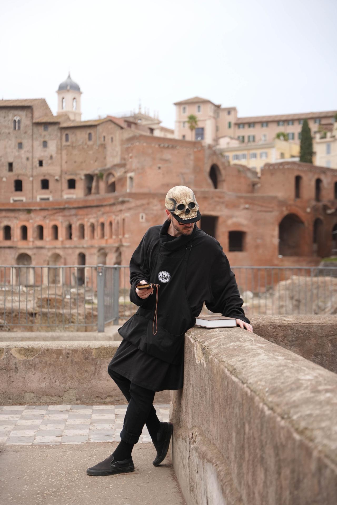 A man in a skeleton baseball hat looks at a book on a wall in front of old buildings