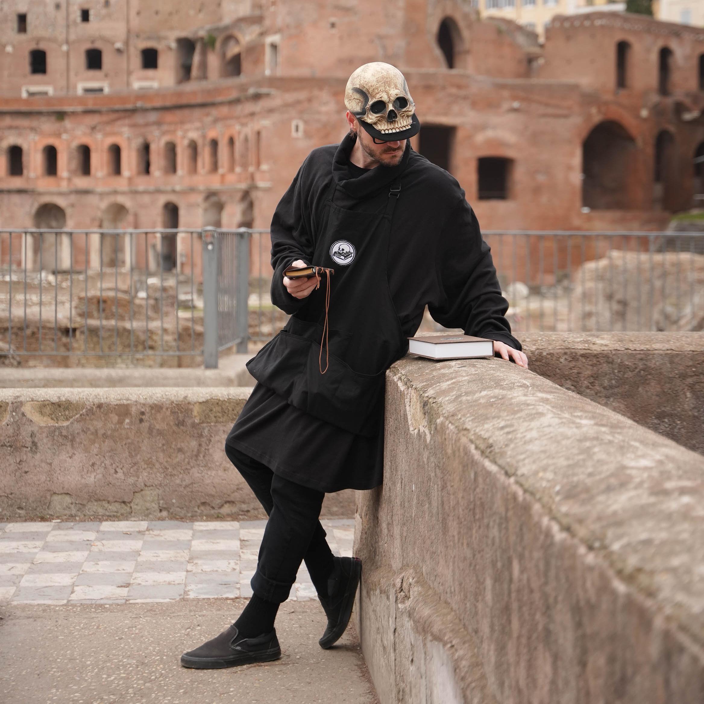 A man in a skeleton baseball hat looks at a book on a wall in front of old buildings