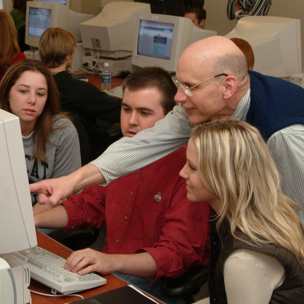 A professor and two students look at an old-style computer monitor. 