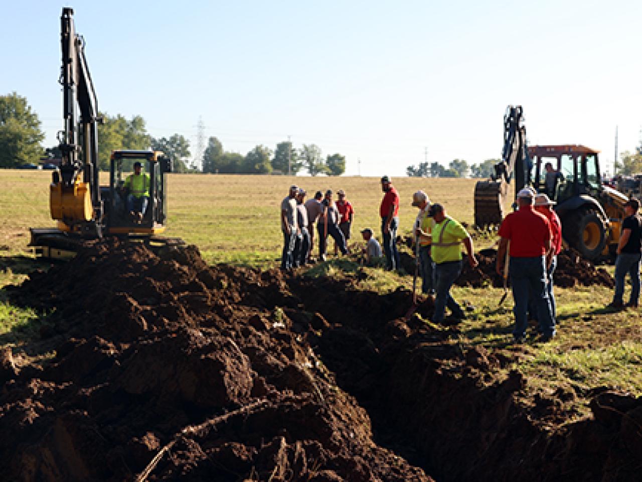 Heavy equipment digs drainage ditches in a field with spectactors looking on