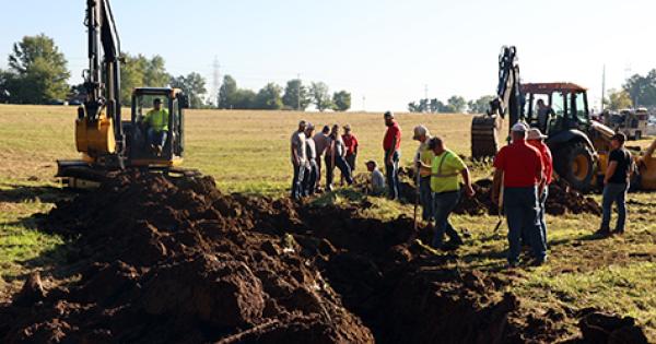 Heavy equipment digs drainage ditches in a field with spectactors looking on
