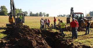 Heavy equipment digs drainage ditches in a field with spectactors lookin on