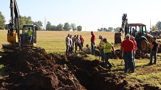 Heavy equipment digs drainage ditches in a field with spectactors lookin on