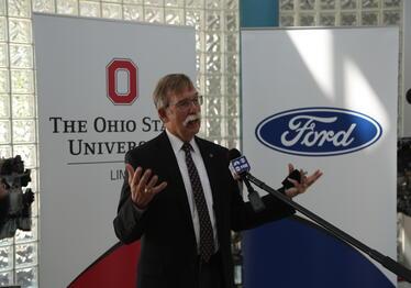 Dean Tim Rehner speaking in front of an Ohio State banner and a Ford motor company banner