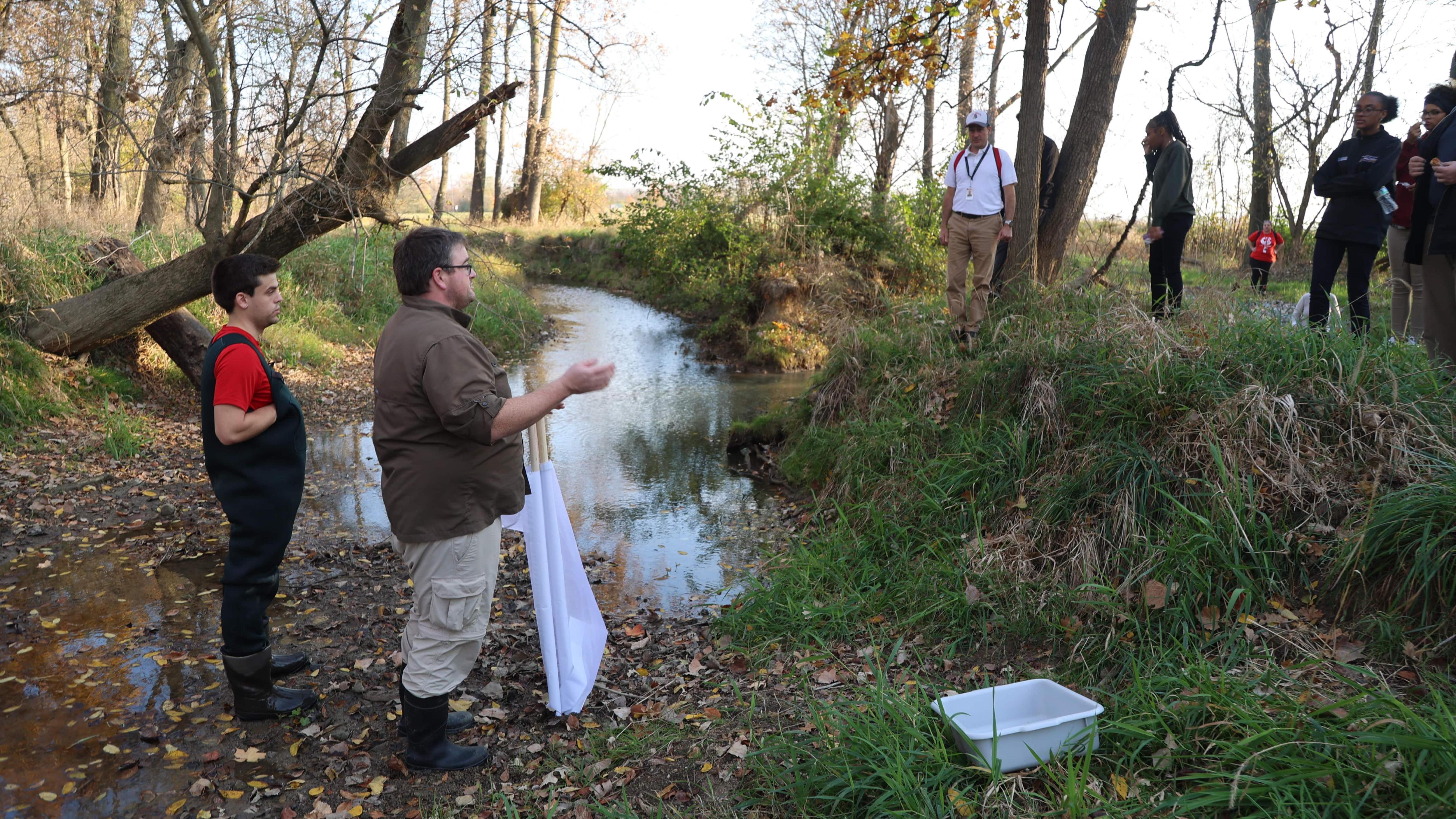 A professor and a student stand in a creek talking to a group on the shore