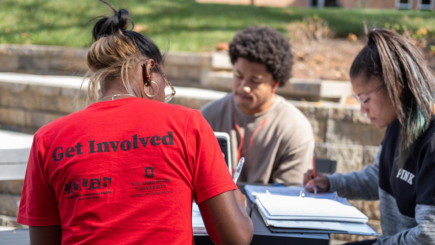 A student with "get involved" on her back studies at an outside table with two other students.