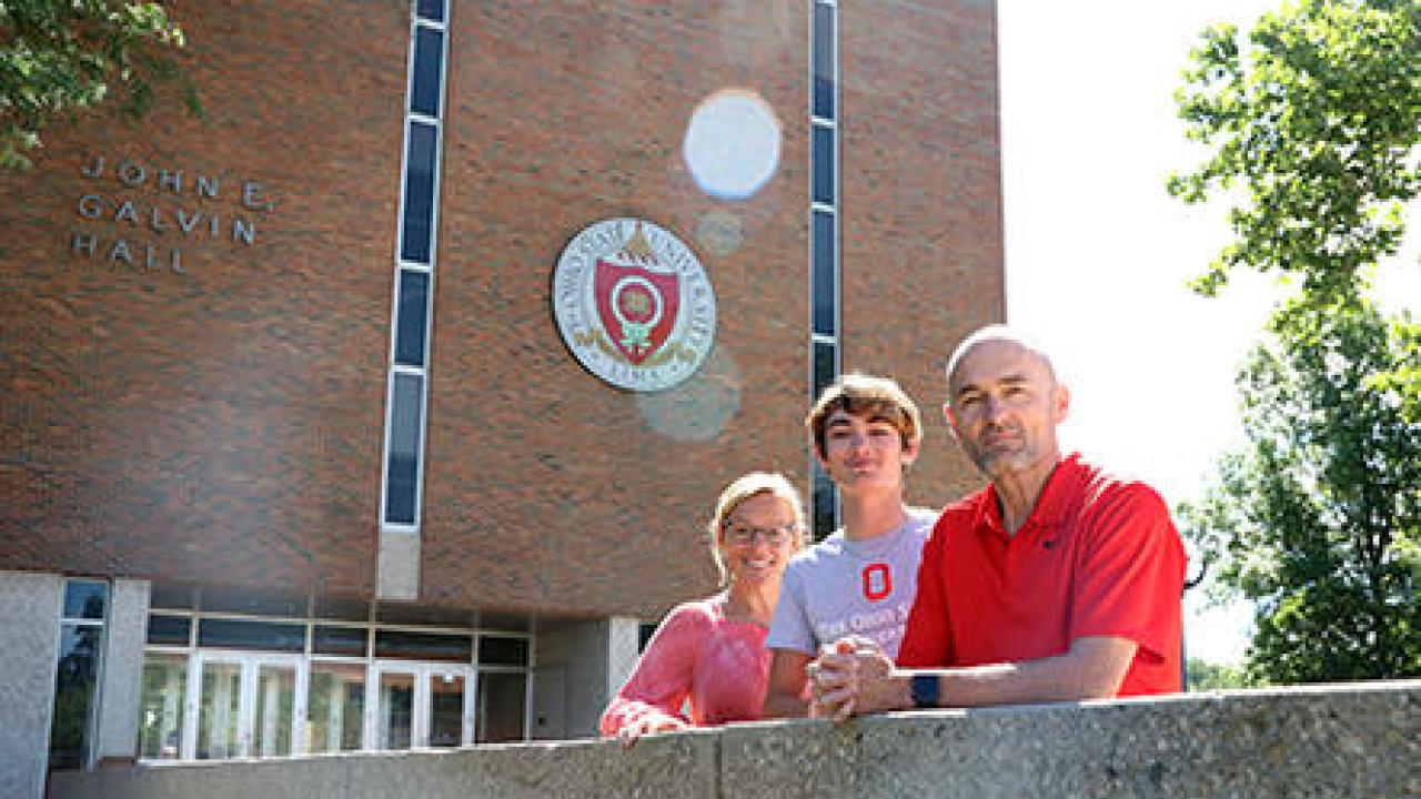 A student with two parents in front of a building on campus